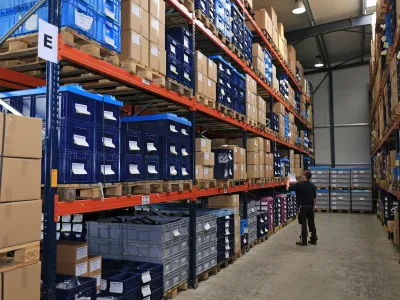 An RH Kunststofftechnik GmbH employee checking a label on plastic containers in the high-bay warehouse, between pallet racks filled with small load carriers.